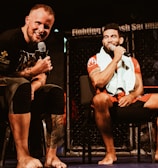 Two individuals are seated on a stage, each holding a microphone and smiling. They are in a sports-related setting with one wearing a casual black outfit and the other in sportswear, with a towel around his neck. The background includes elements of a fighting or martial arts cage.