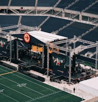 A large outdoor stage is set up inside a stadium, equipped with tall scaffolding, lights, and multiple large display screens. The seats in the stadium are mostly empty. The field is marked with numbers and lines typical of a sports venue. A few people are visible near the stage and on the field.