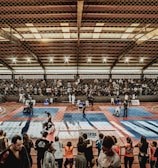 A large indoor arena with a high ceiling, filled with an audience seated on bleachers. In the center, two fighters are engaged in a martial arts competition on blue and red mats. The scene is captured through a mesh fence, adding a textured foreground.