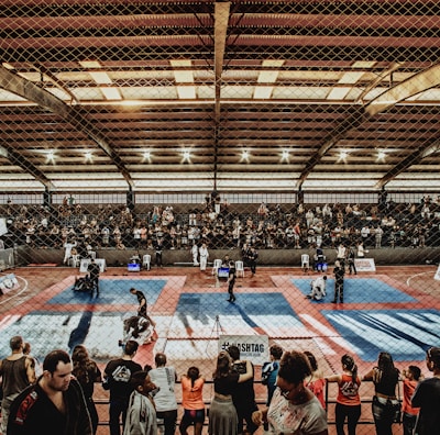 A large indoor arena with a high ceiling, filled with an audience seated on bleachers. In the center, two fighters are engaged in a martial arts competition on blue and red mats. The scene is captured through a mesh fence, adding a textured foreground.