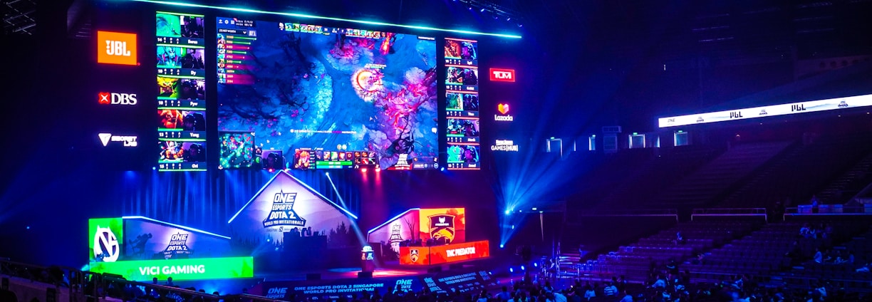 A large esports arena featuring a massive screen displaying a video game, surrounded by sponsor logos and team names. The auditorium is filled with spectators watching the gaming event. Neon blue and purple lights illuminate the stage, creating a vibrant atmosphere.