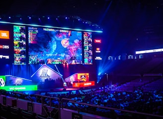 A large esports arena featuring a massive screen displaying a video game, surrounded by sponsor logos and team names. The auditorium is filled with spectators watching the gaming event. Neon blue and purple lights illuminate the stage, creating a vibrant atmosphere.