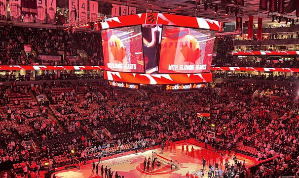 An indoor sports arena filled with spectators and a basketball court illuminated by red lighting. The scoreboards and video screens display promotional content, with numerous banners and championship flags hanging from the ceiling. Players and officials are gathered on the court, possibly in preparation for a game or event.