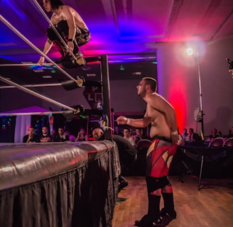 A wrestling match taking place in an indoor venue, where one participant is crouched on the top rope of the ring, while the other stands outside the ring looking at them. A crowd watches the event enthusiastically from their seats.