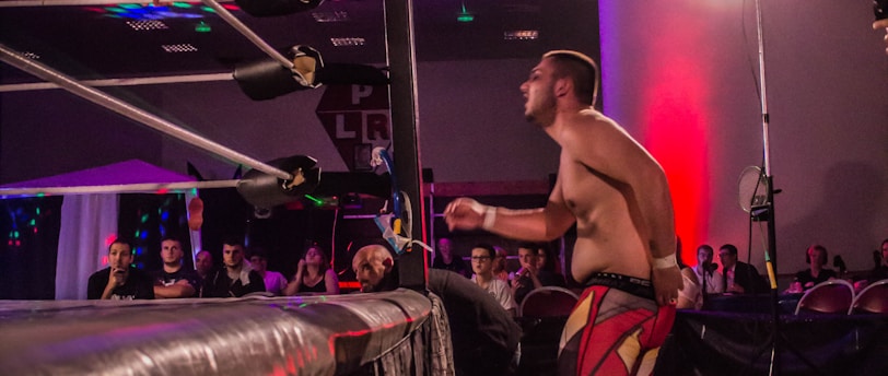 A wrestling match taking place in an indoor venue, where one participant is crouched on the top rope of the ring, while the other stands outside the ring looking at them. A crowd watches the event enthusiastically from their seats.