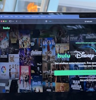 A laptop screen displays a web page featuring a mix of movie and TV show covers, highlighting popular titles and a Hulu and Disney+ bundle promotion. There are visible icons and images of films and shows alongside promotional text offering entertainment and live sports.