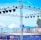 A setup for an outdoor event with a boxing ring surrounded by flags and a metal truss structure equipped with multiple stage lights. The background features a body of water with clear blue skies, and there are white tents on the ground.