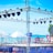 A setup for an outdoor event with a boxing ring surrounded by flags and a metal truss structure equipped with multiple stage lights. The background features a body of water with clear blue skies, and there are white tents on the ground.