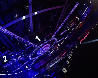 A large indoor arena with colorful and vibrant lighting, featuring a center stage surrounded by rigging and structures. Large numbered screens are visible, indicating marked sections from one to five. Various pieces of equipment and people are scattered around the venue.
