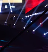 An array of bright stage lights beams across a dark arena, creating a dynamic atmosphere with a large screen displaying various logos and text. A camera crane is visible in the foreground, suggesting a professional event setup.