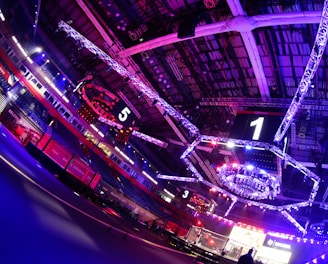 An indoor arena with vibrant purple and blue lighting, featuring large numbered screens suspended from the ceiling. The venue has a high ceiling with intricate metal truss structures and hanging lights, creating a futuristic and energetic atmosphere.
