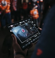 A person is holding a tablet that displays a video or image of a red and blue emblem on the screen. In the background, several people wearing dark and orange colored clothing, possibly sports jerseys, are partially visible. The scene is dimly lit, suggesting an indoor or evening setting.