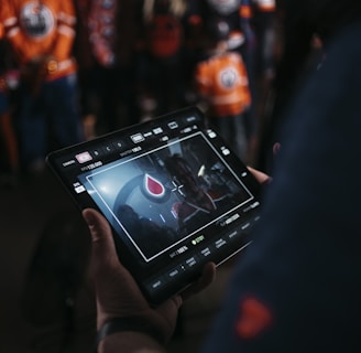 A person is holding a tablet that displays a video or image of a red and blue emblem on the screen. In the background, several people wearing dark and orange colored clothing, possibly sports jerseys, are partially visible. The scene is dimly lit, suggesting an indoor or evening setting.