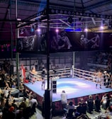 A crowded indoor arena with a boxing ring at the center, surrounded by an audience seated at tables. The scene is illuminated by overhead lights, casting a vibrant glow over the ring where two fighters are preparing for a match. Spectators are engaged, capturing the event with cameras and enjoying the lively atmosphere.