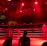 A wrestling ring is illuminated with red lighting, creating a dramatic atmosphere. Several people are visible around the ring, with silhouetted spectators in the foreground. Banners or art adorn the upper walls, and the venue has an industrial ceiling with exposed beams.
