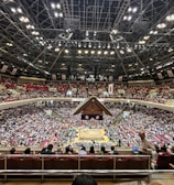 A large indoor arena packed with spectators seated in tiers around a central platform. The platform features traditional Japanese decor, indicating it may be used for sumo wrestling. The atmosphere is bustling, with people filling the seats and many spectators visible in the foreground.
