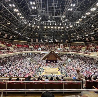 A large indoor arena packed with spectators seated in tiers around a central platform. The platform features traditional Japanese decor, indicating it may be used for sumo wrestling. The atmosphere is bustling, with people filling the seats and many spectators visible in the foreground.