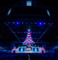 An expansive venue filled with a large audience, featuring a grand stage lit with vibrant blue and pink lights. The ceiling is decorated with a large display, and numerous screens showcase the event's branding. The atmosphere is dynamic and futuristic, reflecting a technologically advanced conference.