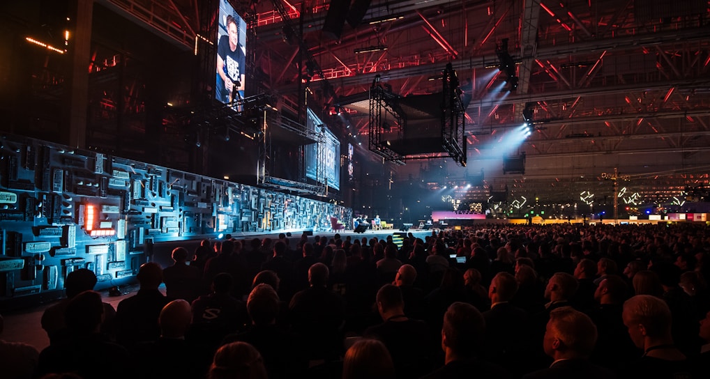 A large indoor event with a stage featuring bright screens and spotlights, attended by a substantial crowd. The venue has a high ceiling with industrial beams and red lighting accents. The atmosphere resembles a concert or a conference with a focus on visual effects.
