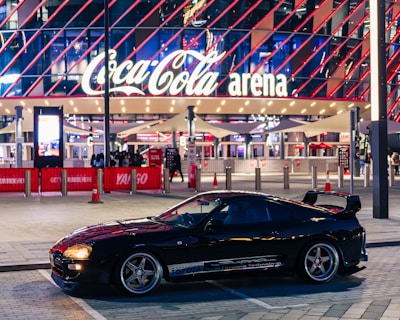 A sleek black sports car is parked in front of an illuminated arena entrance, with a large sign bearing the name of a famous beverage brand. The building features a modern, geometric facade with bright red and blue lights, creating a vibrant and lively atmosphere.